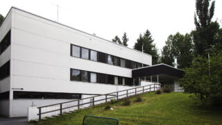 Fi: Moderni, valkoinen rakennus, jossa on suuria ikkunoita ja vihreä nurmikko edessä. Taustalla näkyy puita ja pensaita. / Sv: Modern byggnad i vitt med stora fönster och grön gräsmatta framför. Träd och buskar syns i bakgrunden.