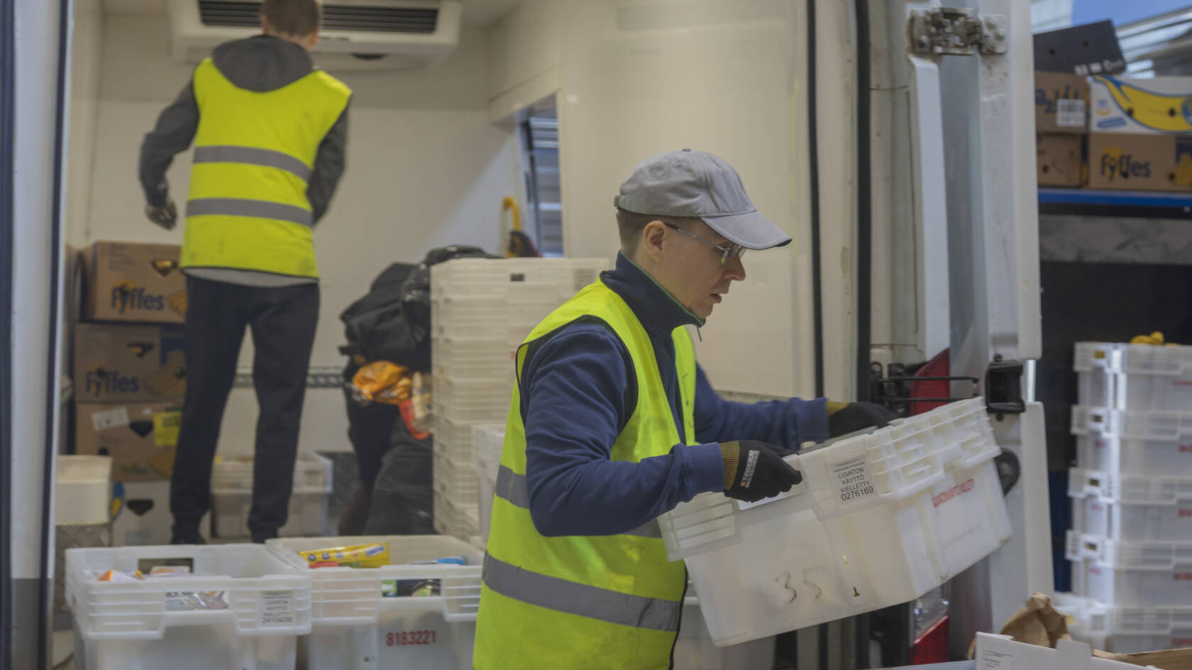 Kaksi henkilöä työskentelee pakettiautossa, jossa on valkoisia laatikoita. Toinen henkilö lastaa laatikoita, toinen järjestää tavaroita.