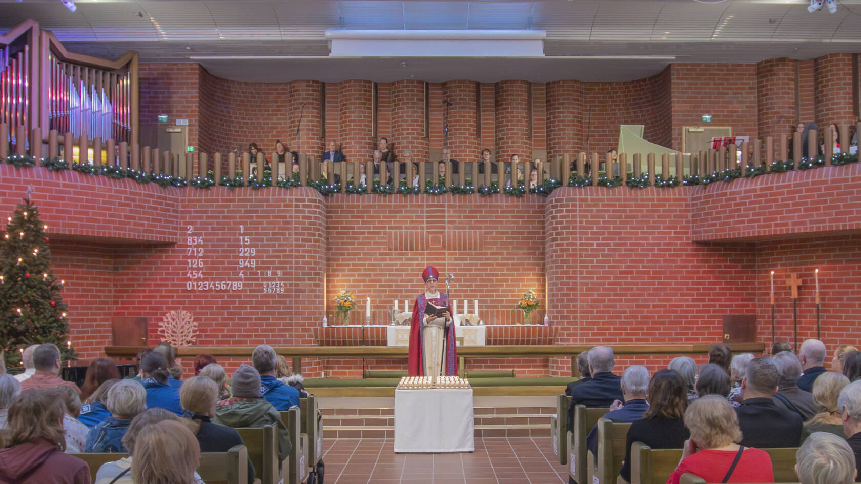 Tiilisen avaran sisätilan keskellä pöydän takana seisoo henkilö liturgisissa vaatteissa. Molemmin puolin istuu ihmisiä penkeissä. Kuvan reunassa on joulukuusi.
