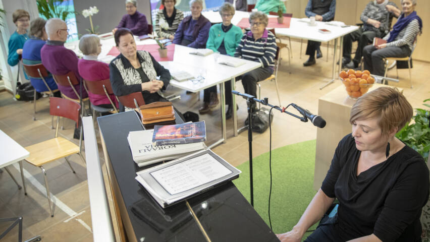 Pianonsoittaja esiintyy ryhmälle vanhempia naisia, jotka istuvat ympäri huonetta.