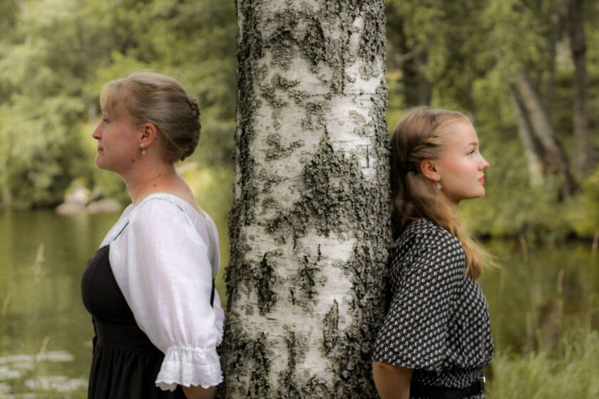 Näkymätön duo kesäisessä maisemassa. Kaksi naista nojaavat saman koivun runkoon katsoen eri suuntiin