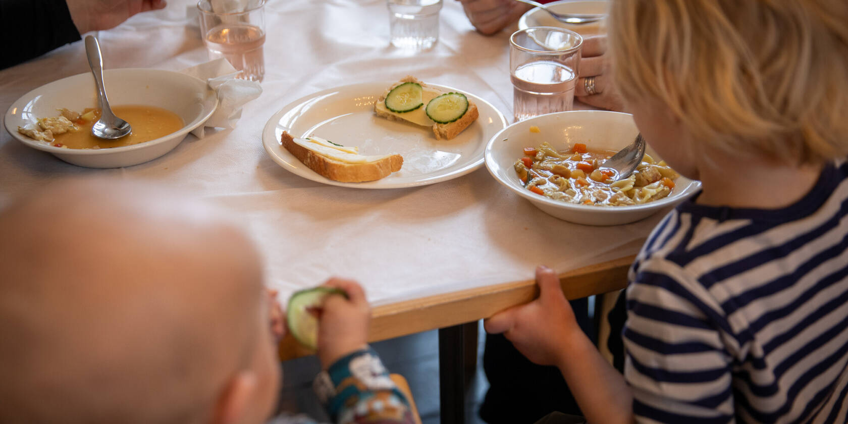 Ruokapöytä, jossa lapset nauttivat ateriaa. Lautasilla on keittoa, salaattia ja leipää. Taustalla näkyy aikuisia ja juomalaseja.