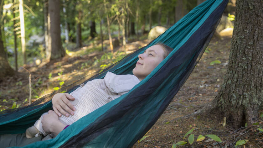 Nuori nainen makaa riippumatossa metsässä, silmät kiinni ja rauhallinen ilme ja hänellä on vauva sylissään ja ympärillä on vihreitä puita.
