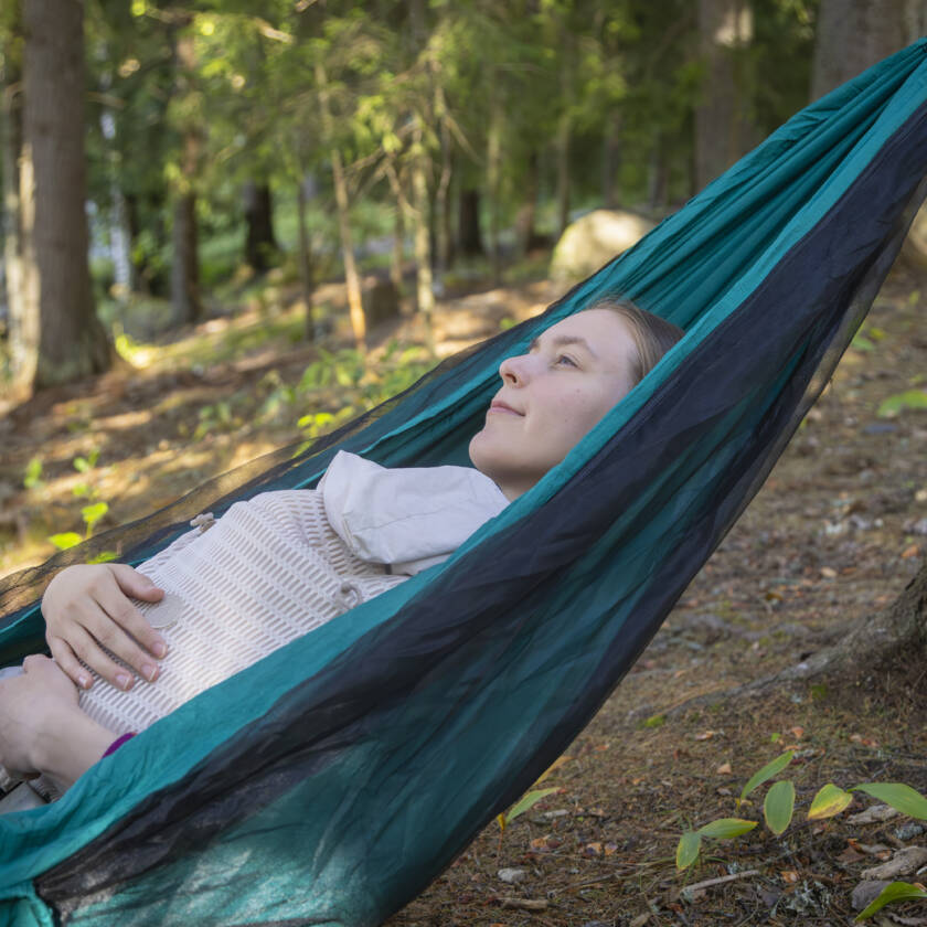 Nuori nainen makaa riippumatossa metsässä, silmät kiinni ja rauhallinen ilme ja hänellä on vauva sylissään ja ympärillä on vihreitä puita.