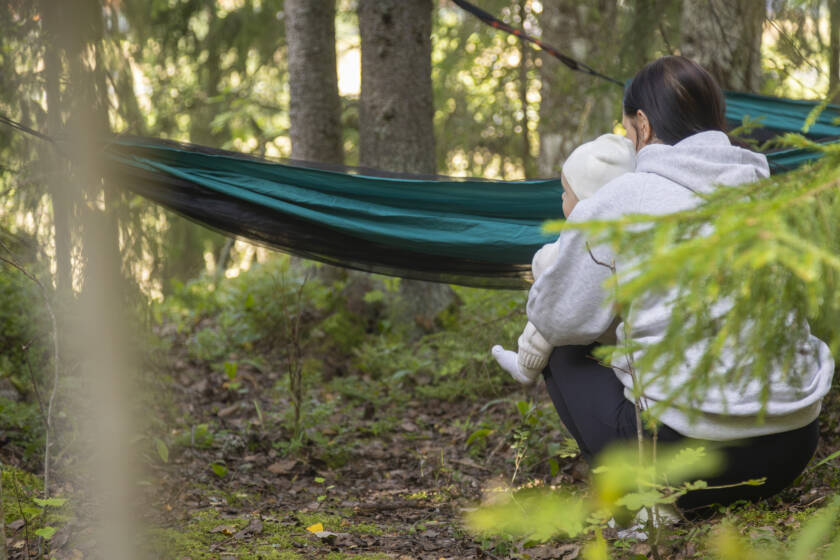 ti kyykyssä metsässä vauva sylissään, taustalla riippumatto ja ympärillä vihreitä puita ja rauhallinen luonto.