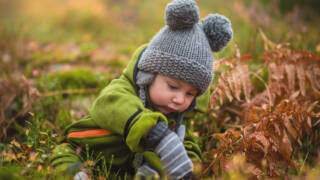 Pieni lapsi leikkii maassa, pukeutuneena vihreään takkiin ja harmaaseen pipoon, tutkien lehtiä ja sammalta syksyisessä metsässä.