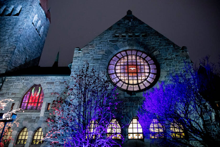 The stone façade of Tampere Cathedral illuminated at night. A large circular stained-glass window glows in the centre, surrounded by blue and purple light.