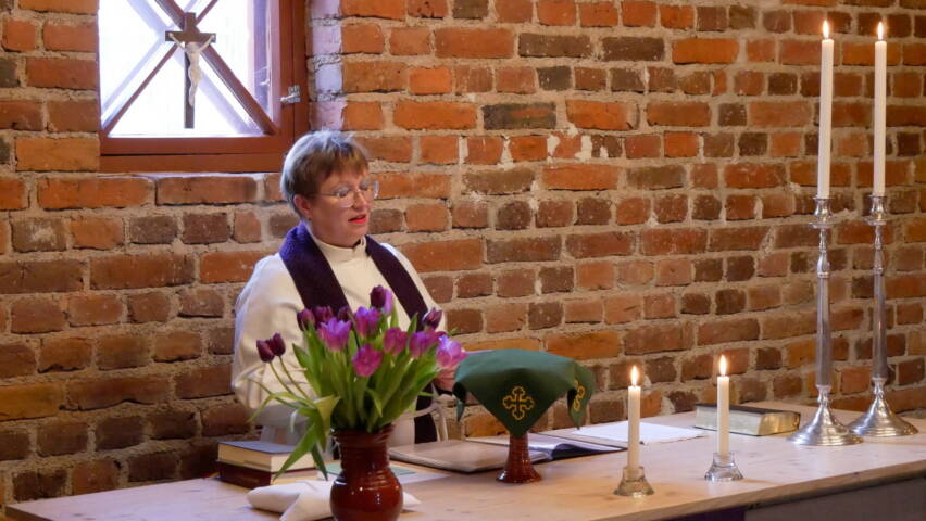 Alttarilla seisova pappi lukee tekstiä kirkossa, pöydällä on Raamattu, palavat kynttilät ja maljakossa violetit tulppaanit, taustalla tiiliseinä ja risti-ikkuna.