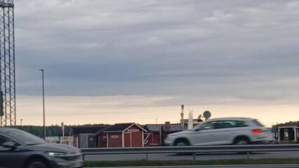 Harmaansinistä taivasta, sen alla oranssinvaaleaa taivasta. Vasemmalla rautaristikkotornia, taivaan alla punaisia mökkejä. Etualalla kahteen suuntaan tiellä ajavat autot.