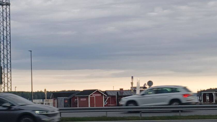 Harmaansinistä taivasta, sen alla oranssinvaaleaa taivasta. Vasemmalla rautaristikkotornia, taivaan alla punaisia mökkejä. Etualalla kahteen suuntaan tiellä ajavat autot.