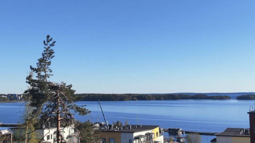 Sininen taivas, järveä, niemeä järven keskellä. Alakulmassa männyn latva ja kerrostalojen kattoja.