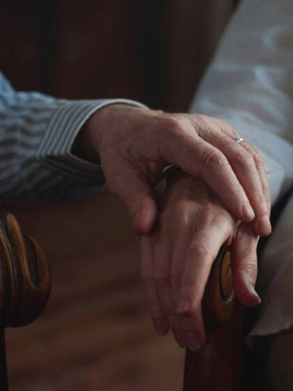 Two hands holding each other sit gracefully on a beautifully carved chair. Dim lighting in the background creates a warm atmosphere.