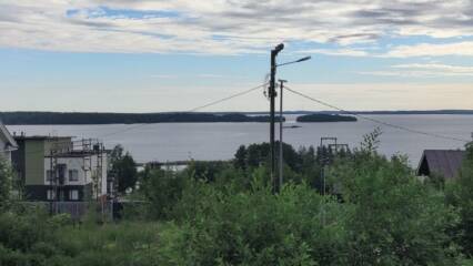 Etualalla pensasta ja nurmikkoa, keskellä pylväs, jonka läpi kulkee sähköjohto, vasemmalla kerrostaloa. Takana järveä, saarta ja pilvistä taivasta.