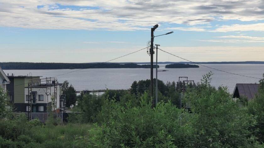 Etualalla pensasta ja nurmikkoa, keskellä pylväs, jonka läpi kulkee sähköjohto, vasemmalla kerrostaloa. Takana järveä, saarta ja pilvistä taivasta.