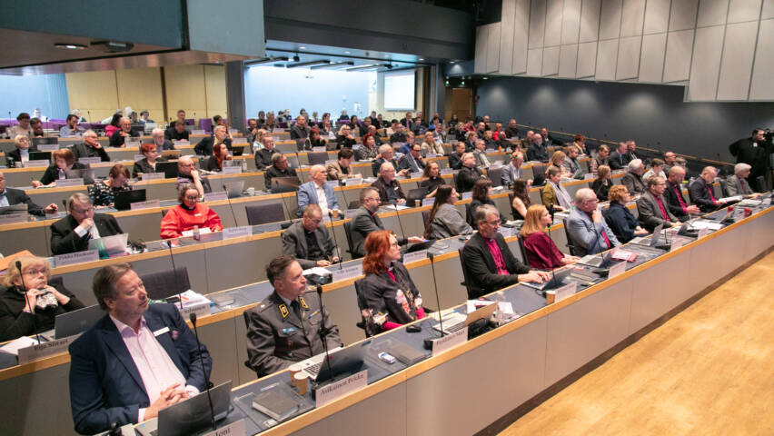 The image shows a large group of people sitting in a hall with tables and computers. They are following a presentation or discussion.
