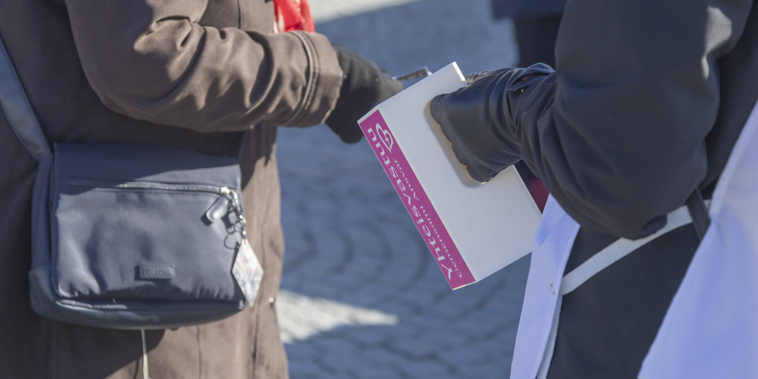 Two people are talking outside; one is holding a white folder, and the other is looking at it. Cobblestones can be seen in the background.