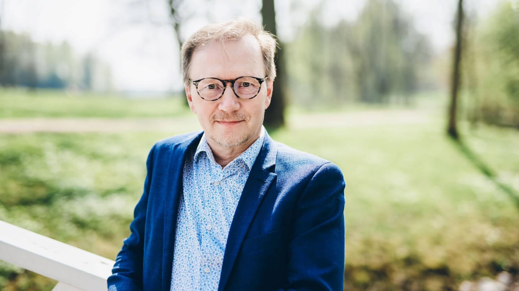 Ylävartalokuva silmälasipäisestä, pikkutakkiin ja paitaan pukeutuneesta miehestä kesäisessä luonnossa.