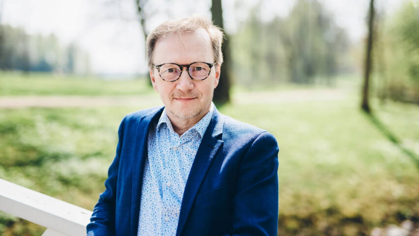 Ylävartalokuva silmälasipäisestä, pikkutakkiin ja paitaan pukeutuneesta miehestä kesäisessä luonnossa.
