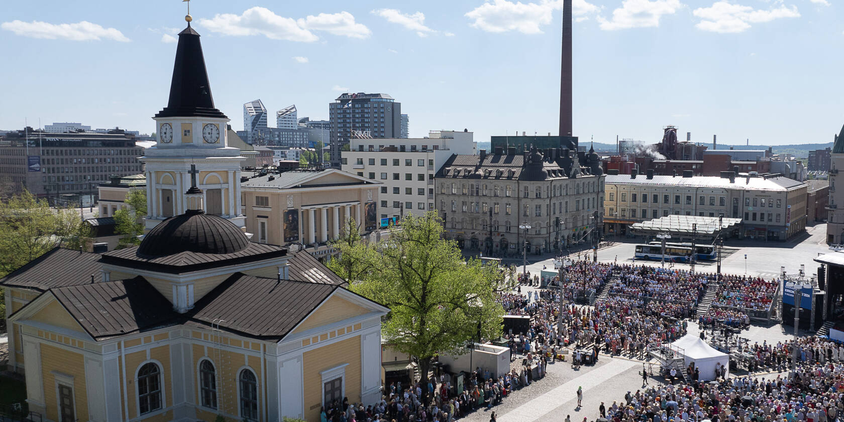 Stadsvy med en stor folkmassa som samlas för ett utomhusevenemang. I bakgrunden en kyrka och moderna byggnader.