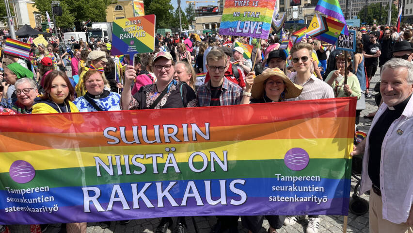 Iso määrä hymyileviä ihmisiä on värikkään raidallisen julistekankaan takana avoimella aukiolla. Takana näkyy kirkon torni ja muita rakennuksia.