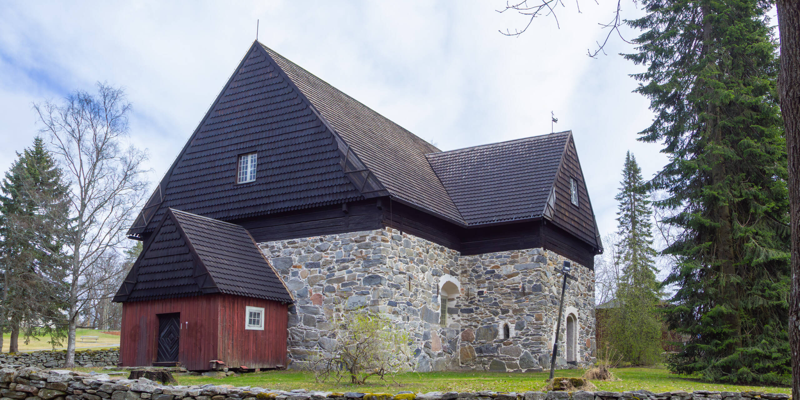 Vanha paanukattoinen kivikirkko, jonka edustalla punainen puinen sisäänkäynti. Taustalla puita.