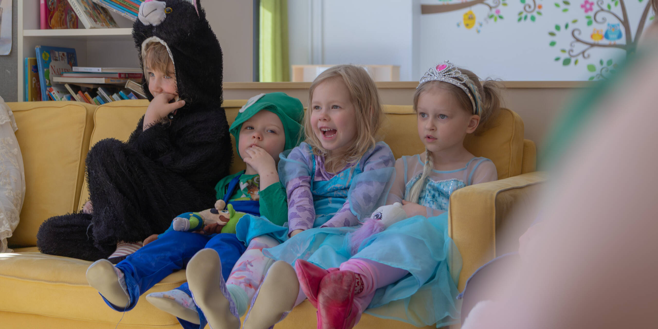 Lapset istuvat värikkäissä vaatteissa keltaisella sohvalla ja nauravat.
