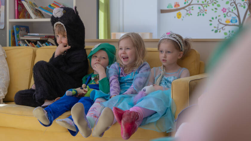 Lapset istuvat värikkäissä vaatteissa keltaisella sohvalla ja nauravat.