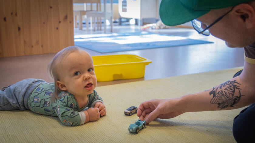 Lapsi makaa mahallaan lattialla ja katsoo kameraan. Isä työntää lelu-puuautoa lapsen edessä matolla.