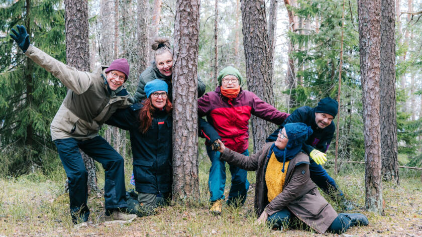 Kuusi ihmistä kuikistelee metsässä puiden takaa levitellen käsiään. Yksi heistä on kontallaan maassa.