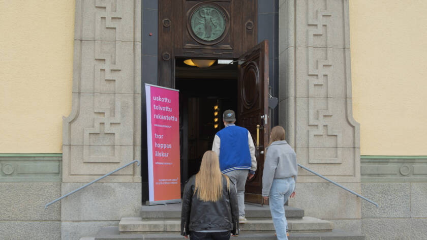 Kolme ihmistä kävelee rakennuksen sisään avoimesta ovesta. Oikealla on kyltti, jossa on tekstiä.