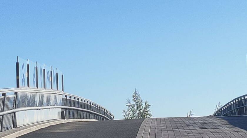 Alareunassa ylöspäin kohoavaa asfalttitietä. Vasemmalla ja oikealla sillan kaiteita. Tien takana koivun latvaa. Sininen taivas.
