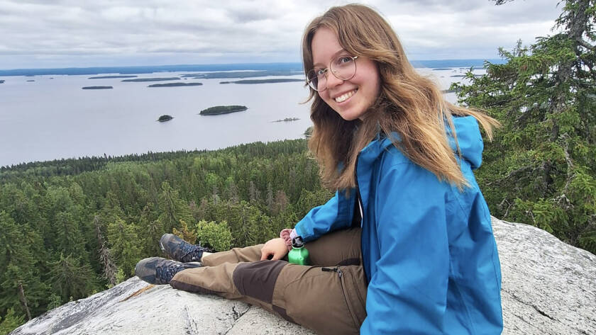 Hymyilevä nainen on kääntynyt katsojaan päin. Hän istuu korkealla kalliolla. Alhaalla on järvinäköala.