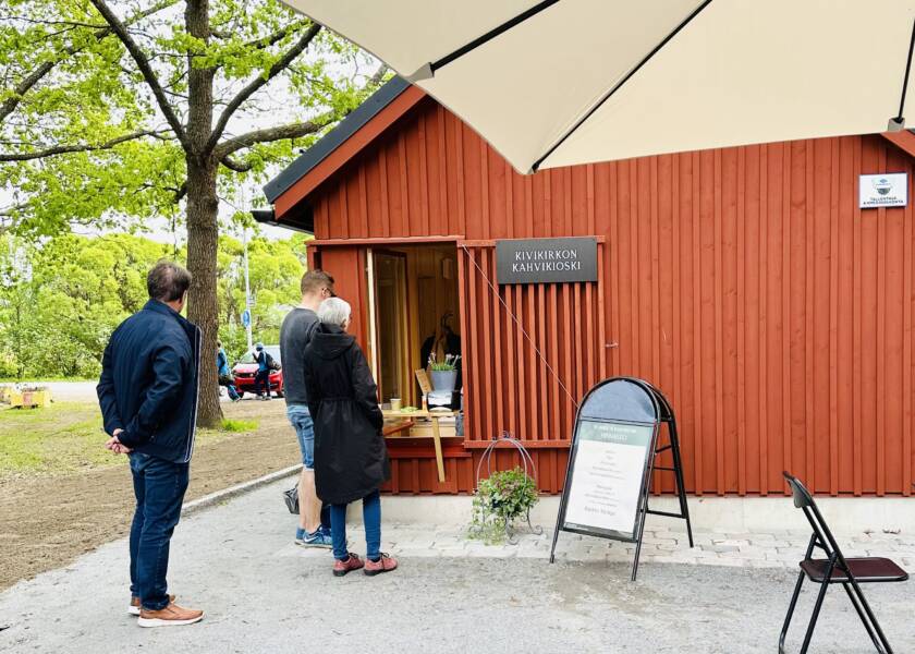 Kolme ihmistä jonottaa punaisen rakennuksen edustalla, jonka taustalla on vihreitä puita.