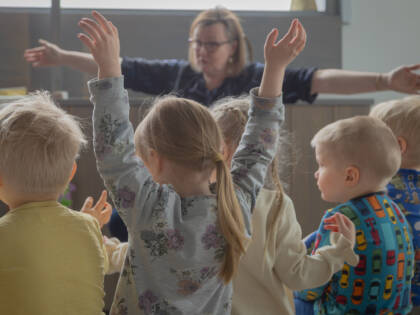 Lapsia istuu penkillä selin kameraan. Yksi lapsista nostaa kädet ilmaan.