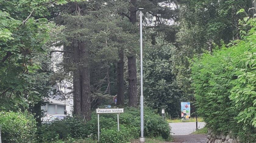 Havu-ja lehtipuita sekä pensaita. Puiden takana rakennusta. Etualalla Pispalan kirkko -kyltti, pylväs ja tie parkkipaikalle, jossa värikäs sähkökaappi.