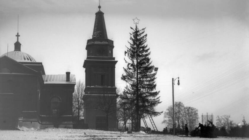 Tumman puukirkon ja kellotapulin vieressä on muotopuoli harva puu, jonka latvassa on tähti. Torin reunassa on ihmisiä tekemässä jotain.