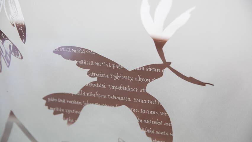 Lasikoristelussa kuva kyyhkystä, sekä Isä meidän -rukouksen tekstiä.