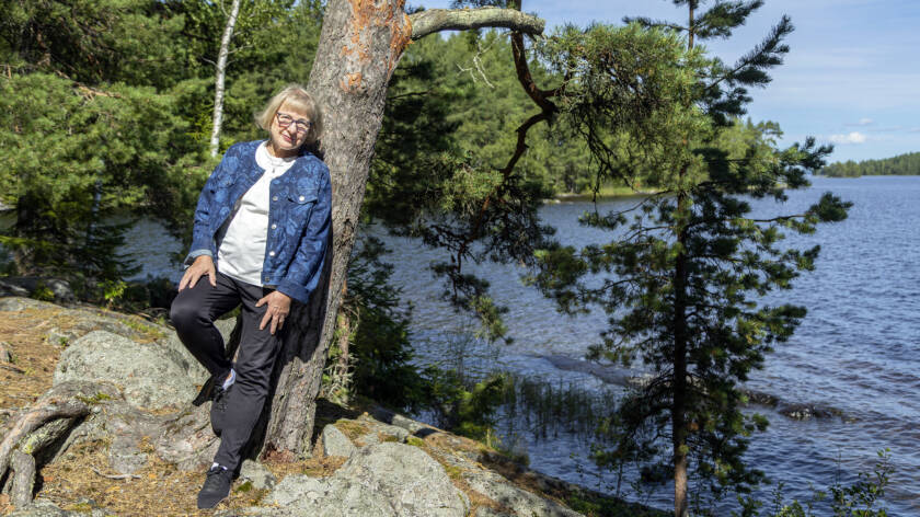 Henkilö nojautuu puun runkoon järven rannalla. Sää on aurinkoinen.