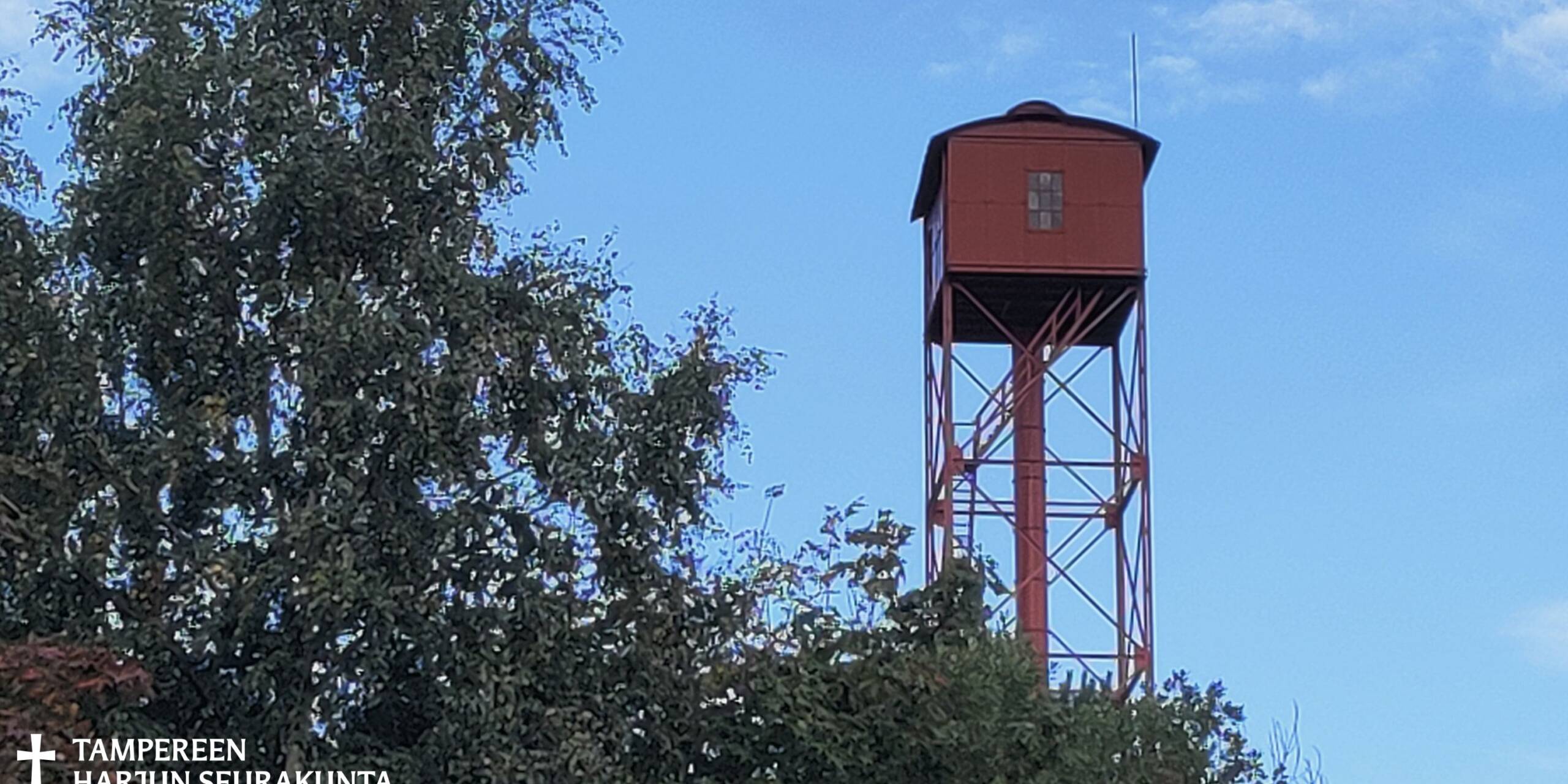 Pieni punainen mökki rakennustornin päällä. Koivun latvaa vieressä, sininen taivas.