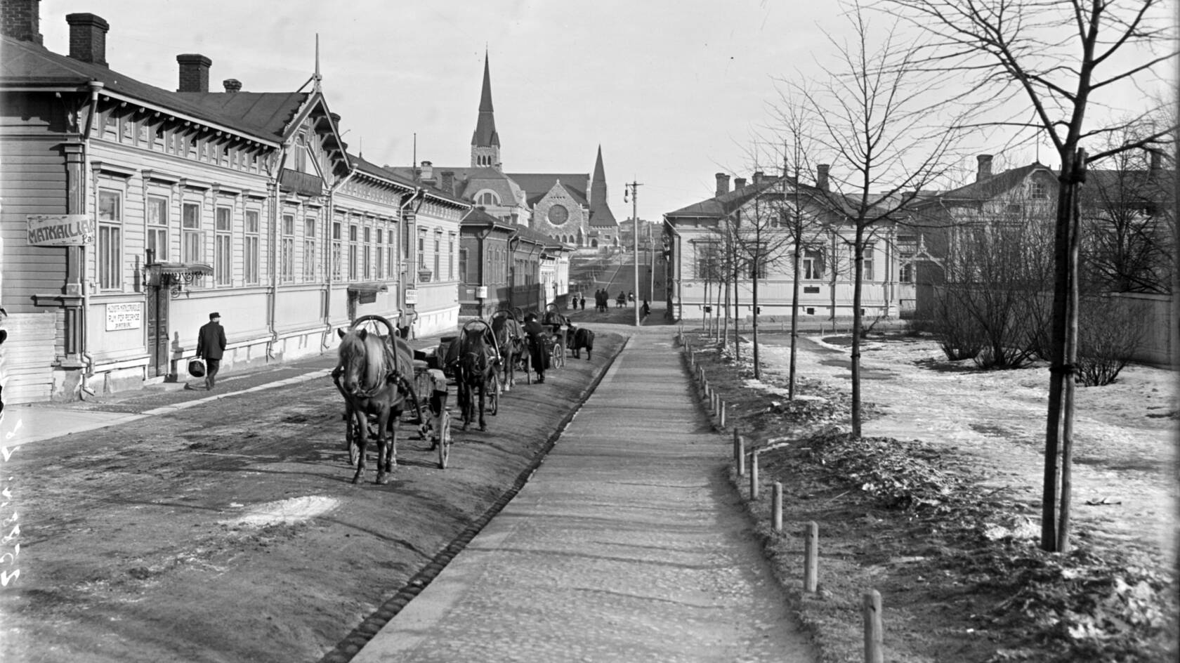 Vanhassa kuvassa vasemmalla puolella kulkee puurakennusten edessä hevoskärryjä multaisella tiellä. Oikealla on pieniä puuntaimia ja takana kirkkorakennus sekä iso puutalo.