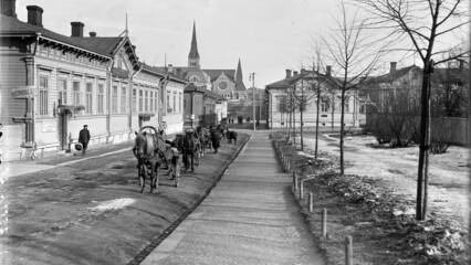 Vanhassa kuvassa vasemmalla puolella kulkee puurakennusten edessä hevoskärryjä multaisella tiellä. Oikealla on pieniä puuntaimia ja takana kirkkorakennus sekä iso puutalo.