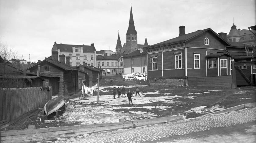 Aukean tontin vasemmalla puolella on kumollaan oleva vene, hökkeleitä ja vasemmalla puutalo. Takana on kirkko ja sen lähellä kivitaloja.