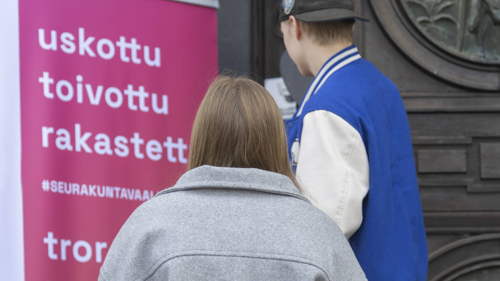 Kaksi henkilöä seisoo kyltin edessä, jossa lukee "uskottu, toivottu, rakastettu", taustalla on puinen ovi.
