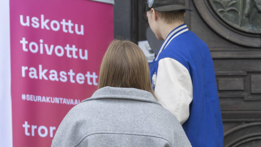 Kaksi henkilöä seisoo kyltin edessä, jossa lukee "uskottu, toivottu, rakastettu", taustalla on puinen ovi.