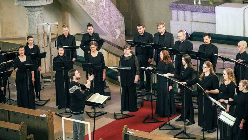 Joukko pitkiin kaapumaisiin vaatteisiin pukeutuneita ihmisiä seisoo sisätilassa kaarimaisessa muodostelmassa ihmisen edessä, joka nostelee käsiään.