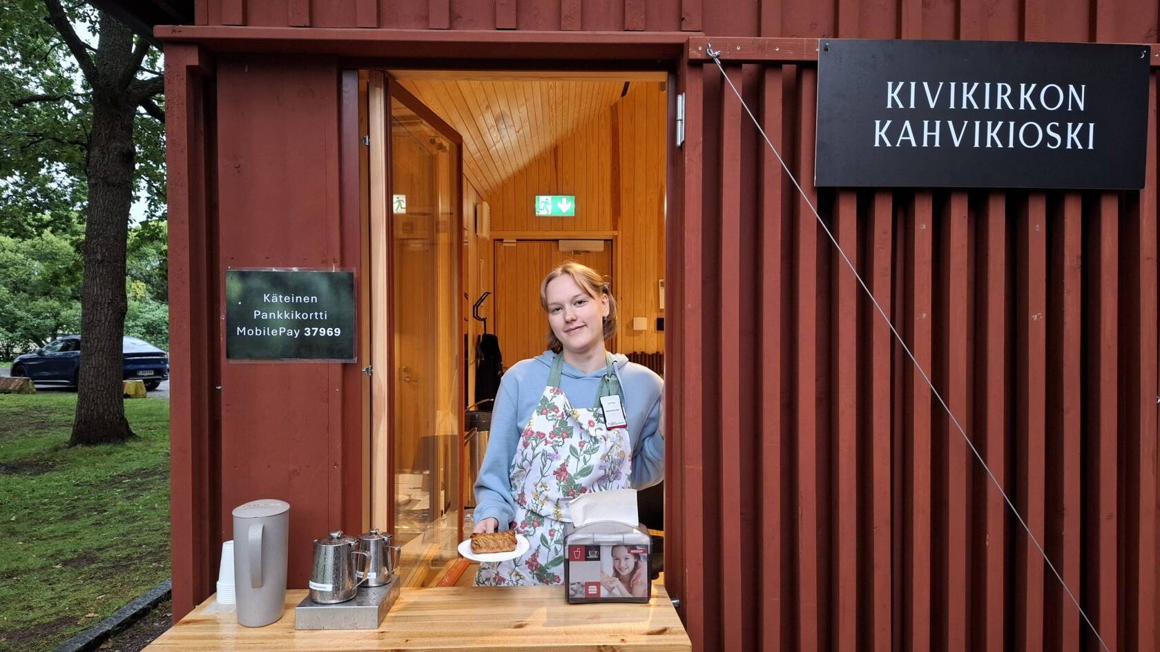 Nuori nainen seisoo pöydän takana oven muotoisen luukun keskellä ja ojentaa lautasella olevaa leivosta kohti katsojaa. Luukku on puisessa rakennuksessa, jonka seinässä olevassa kyltissä lukee kivikirkon kahvikioski.