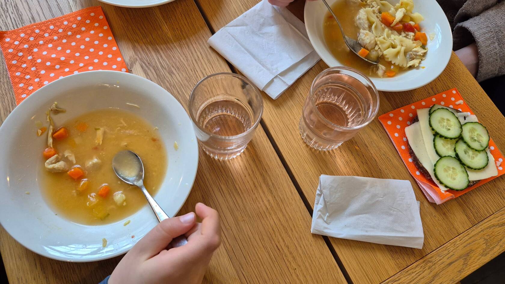 Kuvassa pöydällä lautasia, joilla keittoa. Juomalasit ja leipä. Ihmisistä näkyvät kädet.