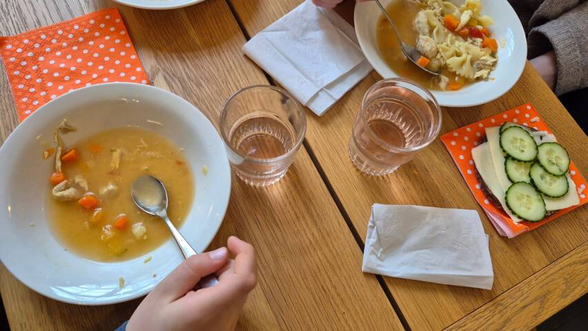 Kuvassa pöydällä lautasia, joilla keittoa. Juomalasit ja leipä. Ihmisistä näkyvät kädet.