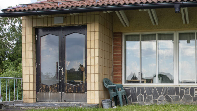 Vaalea laatoitettu rakennus. Tummat pariovet ovat kuluneet. Oikealla pystyjä ikkunaruutuja.