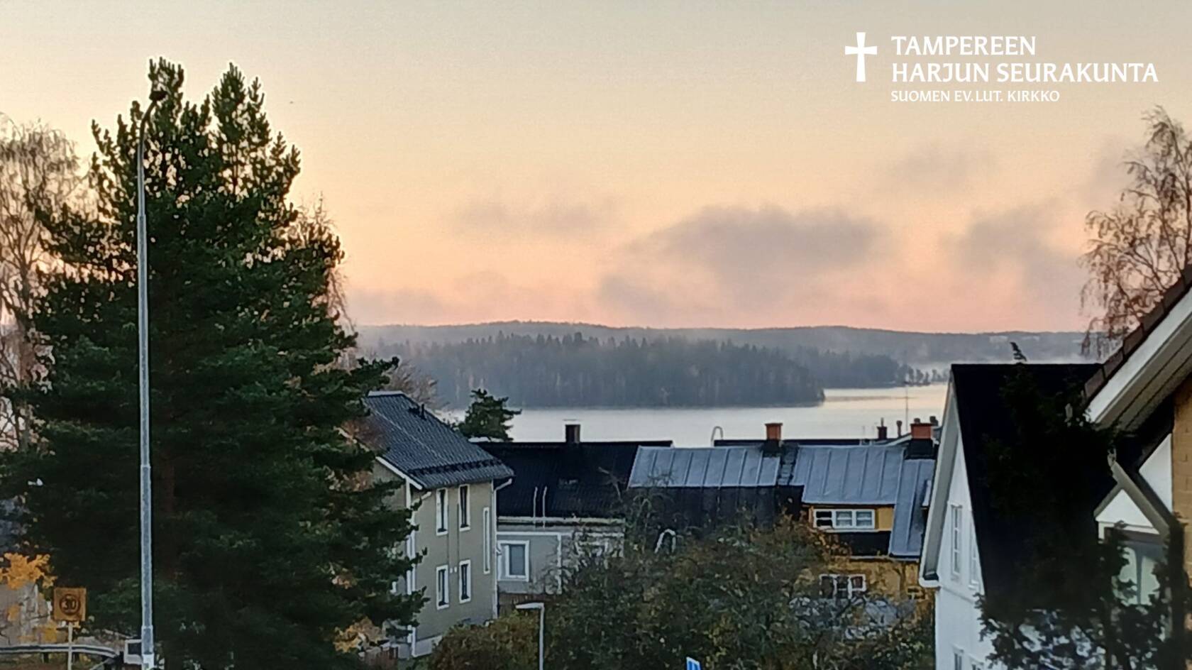 Katu, erivärisiä taloja, puita, järvi ja metsähorisontti. Vaaleanpunainen taivas, sumua tai pilviä ilmassa.